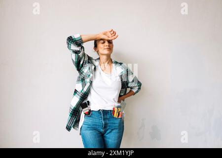 A tired young woman with construction tools in her jeans pockets in a renovated room. An exhausted girl in casual clothes against a white wall. Renova Stock Photo