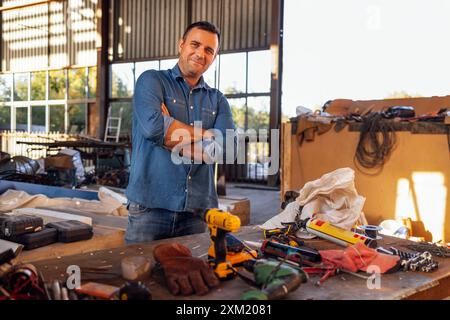A young smiling man in casual clothes stands at a table with construction tools. An attractive builder at a warehouse workplace. A middle-aged enginee Stock Photo