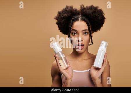 Young Black woman holds two bottles, looking surprised and slightly concerned against a beige background. Stock Photo