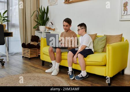 Two boys, one with Down syndrome, are sitting on a yellow sofa at home, using a laptop. Stock Photo