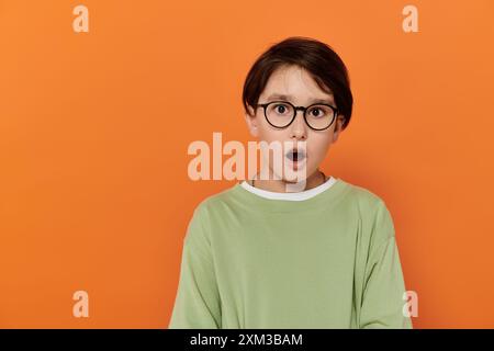 A young boy with glasses looks surprised against an orange backdrop. Stock Photo