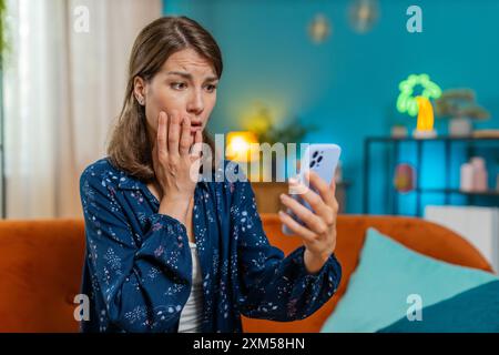 Sad displeased Caucasian woman using smartphone typing browsing, loses becoming surprised sudden lottery game results, bad news, fortune loss fail, deadline, virus. Young girl at home room on couch. Stock Photo