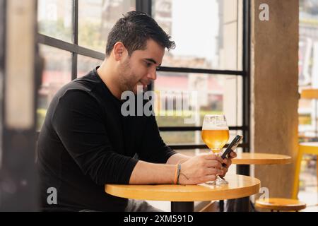 Profile of Latin man sitting enjoying a glass of beer while texting with his smartphone in a pub during the day. The relaxed daytime atmosphere and mo Stock Photo