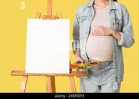 Young pregnant artist holding her belly near canvas on yellow background. Closeup Stock Photo