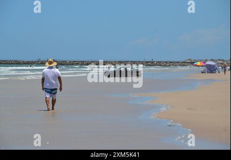 A beachgoer who parked his car too close to the ocean's edge found himself stuck, tires spinning and sand flying, as onlookers watch the scene. Stock Photo