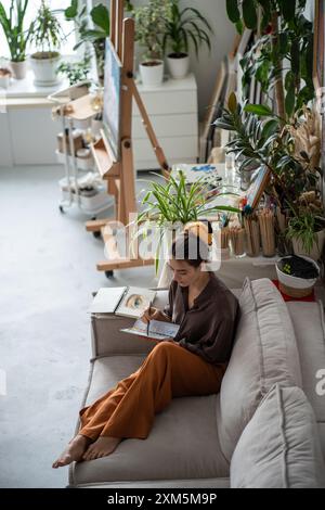 Calm peaceful female artist on couch with sketchbook. Resting woman drawing painting at home studio Stock Photo