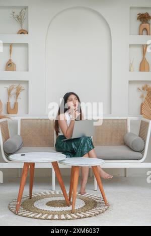 Thoughtful young woman using laptop while sitting on a sofa focused and in deep thought, working on holiday Stock Photo