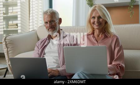 Happy Caucasian elderly married couple senior man woman retired businesspeople business work on Internet together in living room using laptops smiling Stock Photo
