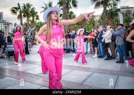 Vibrant outdoor dance Barbie performance, lead dancer in pink, bedazzled outfit strikes a pose with audience gathered amidst palm trees under a cloudy Stock Photo