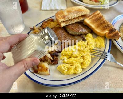 Hand sprinkling salt over breakfast plate with eggs, sausage, toast Stock Photo