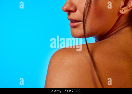 Close-up side view of woman's face and shoulder, highlighting her smooth, radiant skin and healthy complexion in neon light against blue background. Stock Photo
