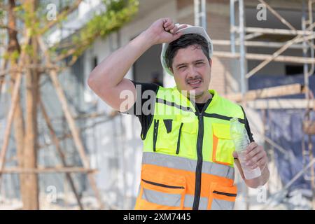 Construction worker in a hard hat and safety vest drinking water, staying hydrated during work on a building site. Stock Photo