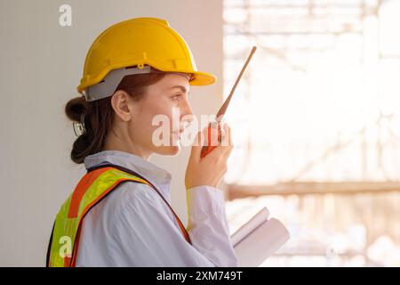 Female construction worker wearing safety gear and hard hat, communicating using a radio, and holding blueprints at a construction site. Stock Photo