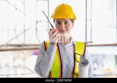 Female construction worker wearing safety gear and hard hat, communicating using a radio, and holding blueprints at a construction site. Stock Photo