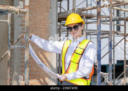 Confident female architect in safety gear holding blueprints and inspecting construction site, demonstrating professionalism and project management. Stock Photo