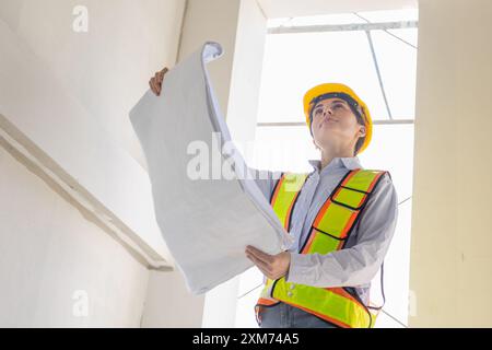 Confident female architect in safety gear holding blueprints and inspecting construction site, demonstrating professionalism and project management. Stock Photo