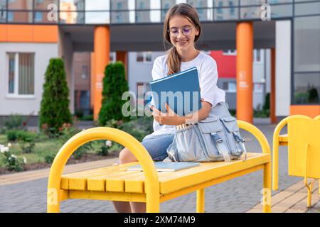 A girl with a backpack near the school and a blue notebook. On the background of the school. Stock Photo