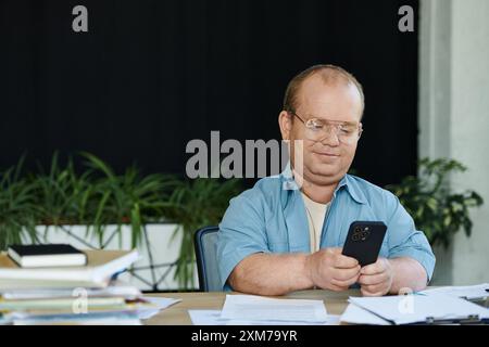A man with inclusivity wearing glasses looks at his smartphone while sitting at a desk. Stock Photo