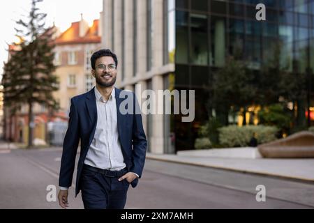 Confident young businessman wearing glasses walking outdoors in city. Smiling professional in jacket and shirt enjoying urban environment. Modern office building in background. Stock Photo