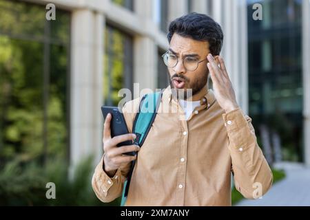 Young man in glasses standing outside looking shocked while checking his smartphone, expressing surprise and disbelief. Professional setting with modern building and greenery in background. Stock Photo