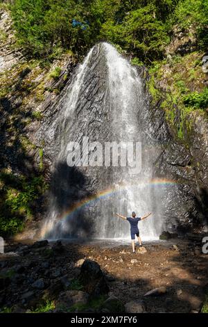 Rear view of young man standing with arms raised in front of the cascade du Queureuilh waterfall. A beautiful rainbow appears in the waterfall. Stock Photo
