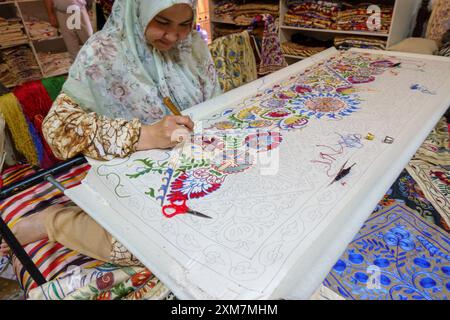 Uzbek woman meticulously hand-embroidering colorful floral patterns on a large fabric using a needle in a textile shop Stock Photo