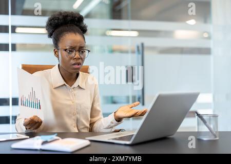 Frustrated businesswoman analyzing financial report while sitting at office desk with laptop. Professional woman experiencing work stress or confusion in modern corporate environment Stock Photo