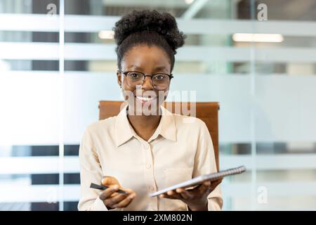 Confident businesswoman smiling during office video call. Professional holding notepad pen, ready for meeting. Modern office setting enhances corporate ambiance. Concept of remote work communication Stock Photo