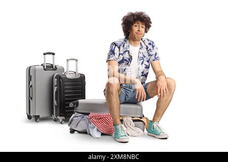 Tired young man sitting on a suitcase full of clothes isolated on white background Stock Photo