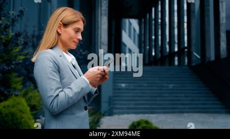 Focused successful businesswoman walking outdoors holding smartphone writing email message communicating with friend or client on social media using Stock Photo