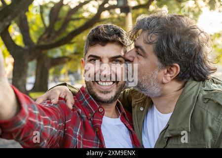 A happily married gay couple is smiling and kissing while taking selfies. Stock Photo