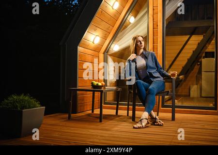 Young woman sitting comfortably on a wooden porch, surrounded by the warm glow of house lights. Concept of serene and tranquil vacations at log cabin Stock Photo