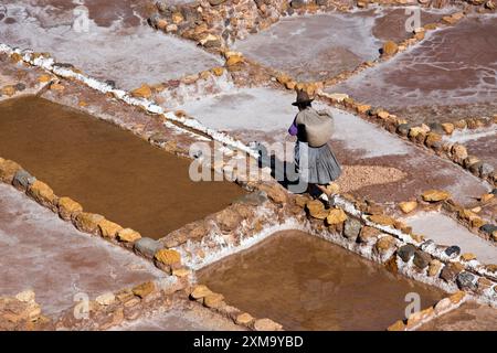 Local woman working at the Maras Salt Mines, located on a mountainside above Urubamba in Peru. The salt mines consist of numerous shallow pools filled with salt water. As the water evaporates, it leaves behind crystallised salt. These salt pans have been in use for over 500 years and are still operated by local families to extract Peruvian pink salt. Stock Photo