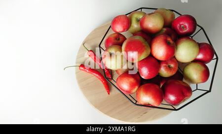 A black geometric bowl filled with red and green apples is placed on a wooden board with three red chili peppers beside it Stock Photo