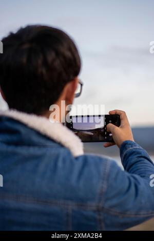 Vertical portrait of a young bearded man with glasses taking a picture of the landscape with his cell phone Stock Photo