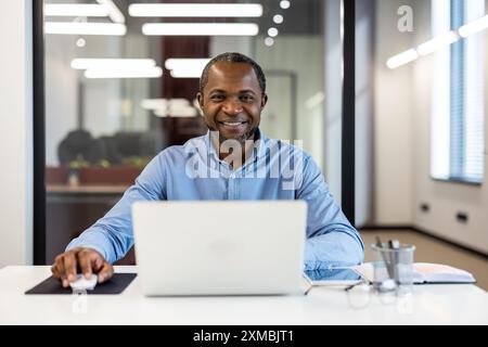 Confident businessman seated at desk smiling while working on laptop in modern office setting. Professional environment with bright lighting and contemporary design. Stock Photo