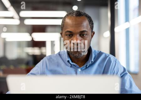 Portrait of focused office worker in blue shirt working on laptop in modern office environment. Depicts concentration, productivity, and professional setting. Suitable for business, technology, and workplace Stock Photo