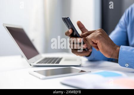 Business professional using smartphone while working on laptop. Modern office with technology. Hands interacting with mobile phone and laptop screen. Concept of remote work, multitasking Stock Photo