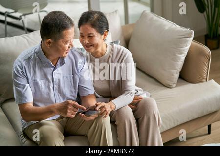 happy senior asian couple sitting on couch at home using cellphone together Stock Photo