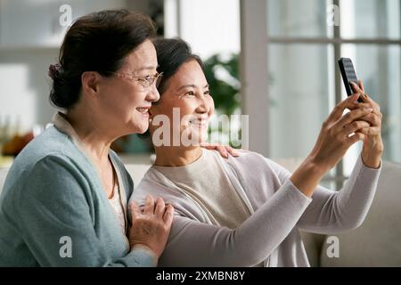 two happy senior asian women sitting on couch at home taking a selfie using cellphone happy and smiling Stock Photo