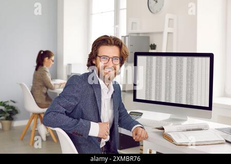 Young bearded Caucasian man data science specialist smiling sits at table in modern office Stock Photo