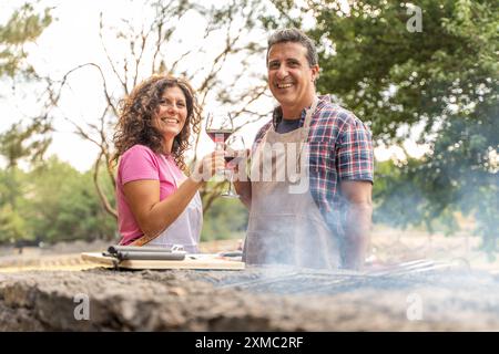 Cheerful middle-aged couple toasting with red wine glasses during a barbecue in nature. Man and woman in aprons sharing a romantic moment while grilli Stock Photo