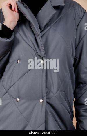 Close-up vertical photo of a grey quilted coat with diamond patterns, showing details of the fabric and buttons, worn by a woman against a beige backg Stock Photo
