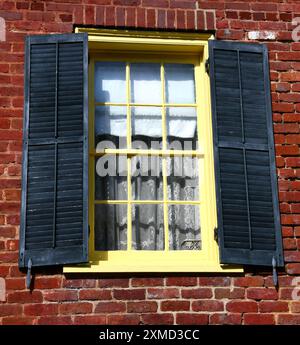 Window, on old, red brick building, is frame with yellow painted wood.  Shutters are black. Stock Photo