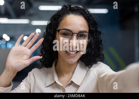 Smiling woman with glasses in office environment having video call. Happy professional waving hand and looking at camera, conveying friendliness and engagement. Modern office setting Stock Photo