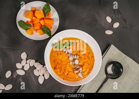 Vegetarian dish: traditional cream soup of pumpkin, carrot with pumpkin seeds in a white bowl and sliced pumpkin on a white plate on a gray background Stock Photo