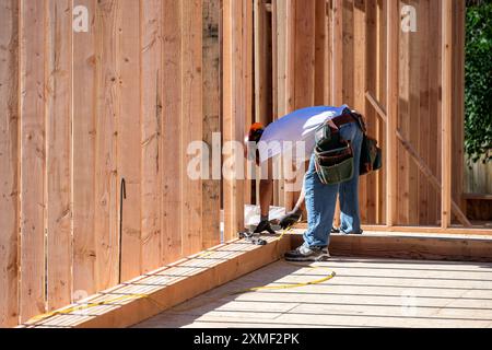 Construction worker in toolbelt and hard hat working with a hammer on new house construction wood framing, summer construction season Stock Photo