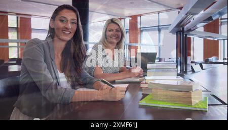 Image of globe and programming language over smiling caucasian teachers sitting in school Stock Photo