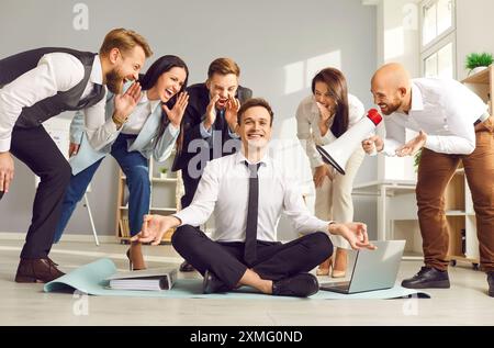 Calm businessman is meditating sitting in office on floor next to his colleagues who are shouting. Stock Photo