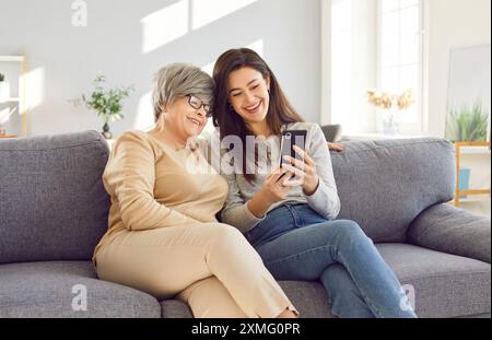 Adult woman using mobile phone with senior mother sitting at home and having online video call. Stock Photo
